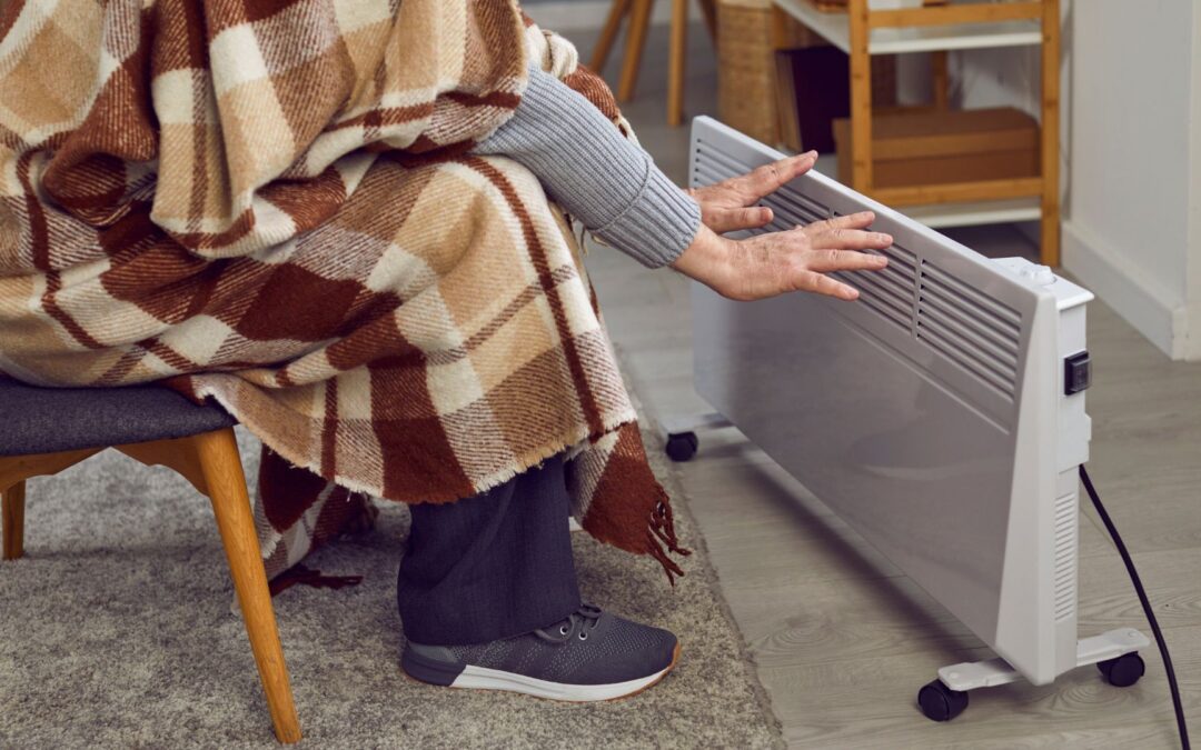 elderly man wrapped in brown plaid flannel blanket seated in front of space heater waving hands to get warmer
